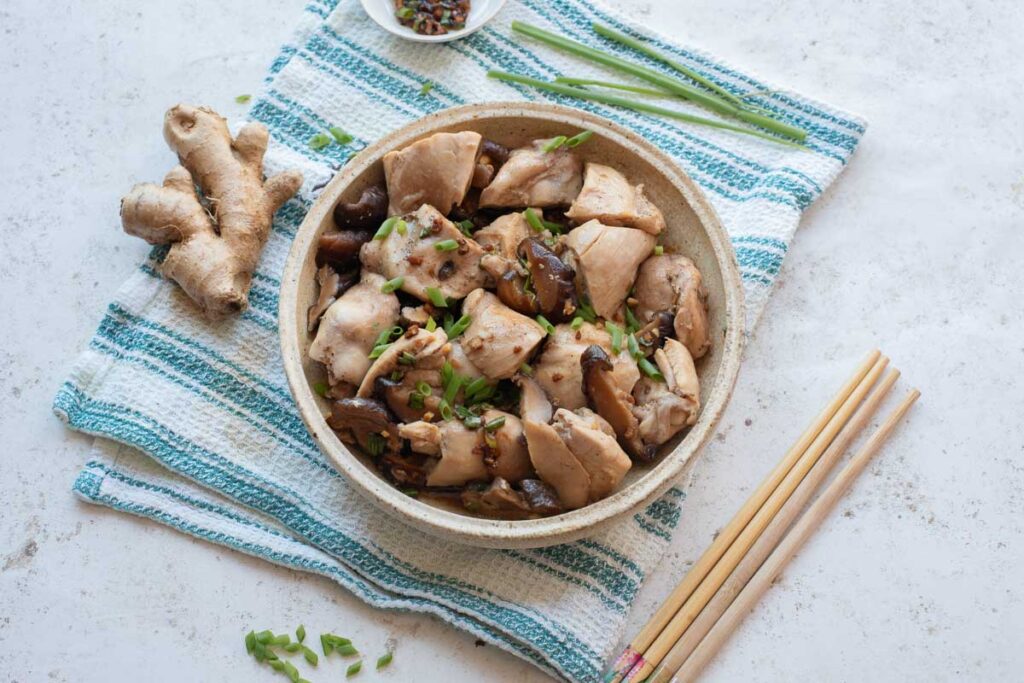 A bowl of Chinese Steamed Chicken with mushrooms and green onions, placed on a striped towel with chopsticks, ginger root, and soy sauce on the side.
