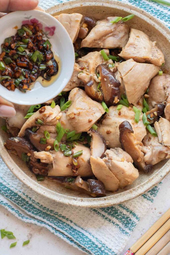 A bowl of Chinese Steamed Chicken with mushrooms and chopped green onions, with a hand holding a small dish of soy-based sauce above the bowl.