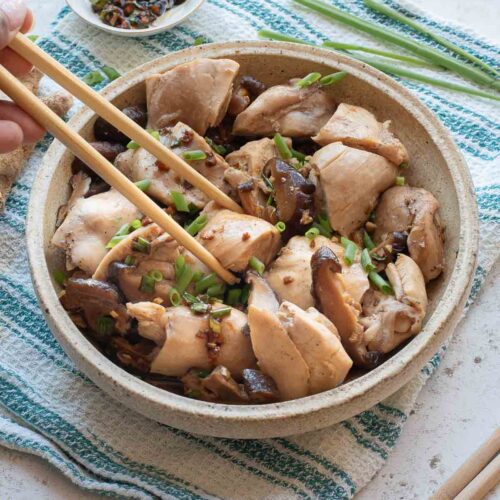 A bowl of Chinese Steamed Chicken pieces garnished with green onions, with chopsticks picking up a piece and a small dish of dipping sauce nearby.