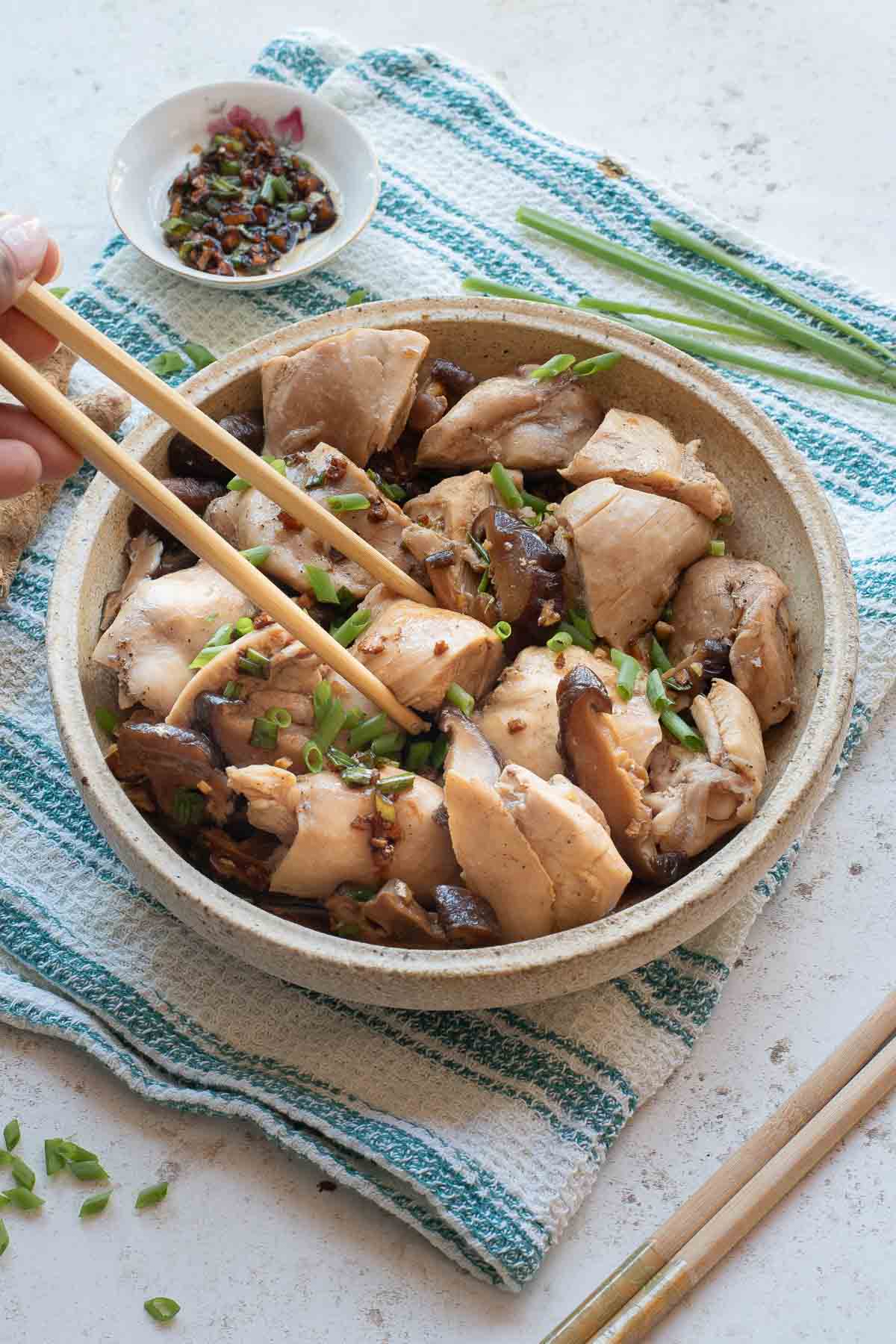 A bowl of Chinese Steamed Chicken pieces garnished with green onions, with chopsticks picking up a piece and a small dish of dipping sauce nearby.