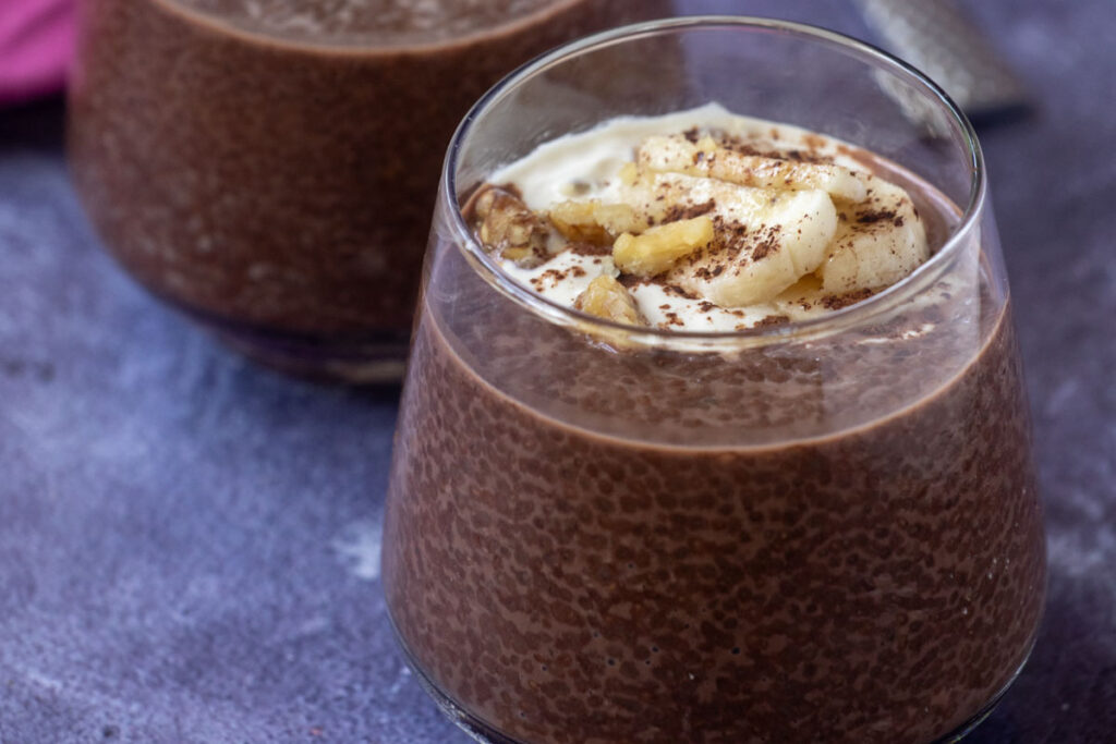 A glass of Chocolate Chia Pudding topped with sliced banana, chopped nuts, and a sprinkle of cocoa powder, set on a purple surface.
