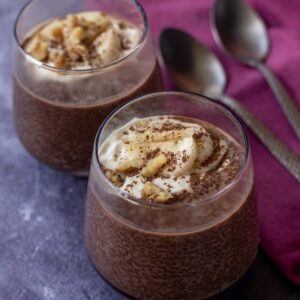 Two glasses of rich Chocolate Chia Pudding topped with banana slices and grated chocolate sit on a purple surface, accompanied by two metal spoons and a folded burgundy napkin.