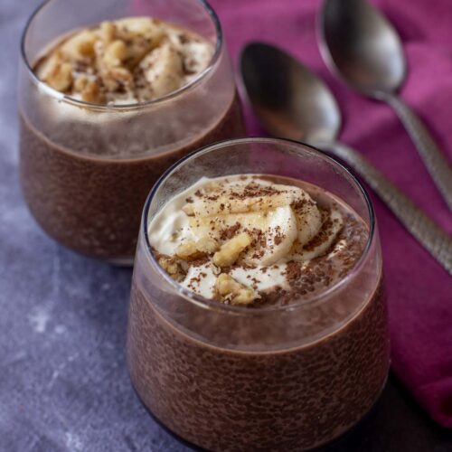 Two glasses of rich Chocolate Chia Pudding topped with banana slices and grated chocolate sit on a purple surface, accompanied by two metal spoons and a folded burgundy napkin.