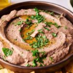 A bowl of Black Bean Hummus topped with chopped parsley and a drizzle of olive oil, surrounded by crispy tortilla chips.