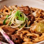 A bowl of Black Bean Noodles stir-fried with beef, cabbage, green onions, and sauce, garnished with shredded scallions and served with chopsticks.