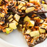 Caramel Apple Bark pieces topped with chopped apples, pretzels, caramel, and a dark chocolate drizzle are served on a white plate.
