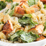 A plate of Chicken Caesar Pasta Salad featuring rotini, grilled chicken chunks, romaine lettuce, croutons, and grated cheese.