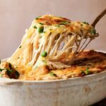 A serving of Chicken Tetrazzini casserole with melted cheese, green peas, and herbs is being lifted from a white dish with a spoon.