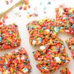 Fruity Pebble Rice Krispies bars with mini marshmallows are cut into squares and arranged on a white surface, with a gold knife and scattered cereal pieces nearby.