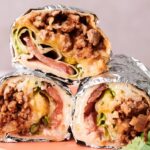 Three halves of foil-wrapped Grilled Cheese Burritos filled with ground meat, melted cheese, lettuce, tomato, and other vegetables are stacked on a plate.