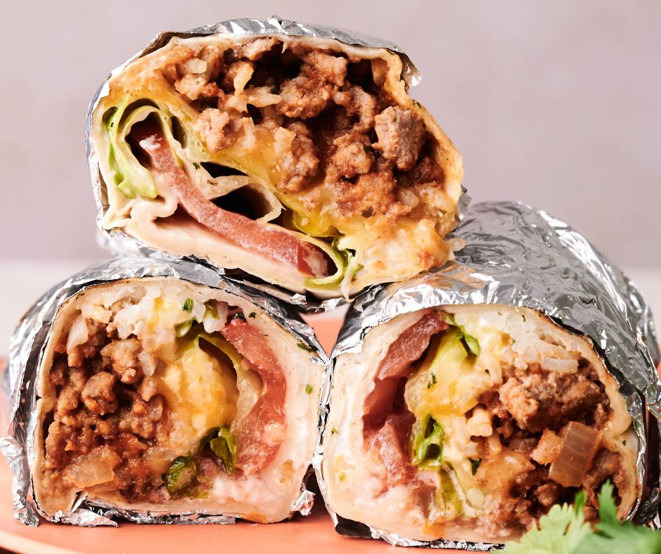Three halves of foil-wrapped Grilled Cheese Burritos filled with ground meat, melted cheese, lettuce, tomato, and other vegetables are stacked on a plate.