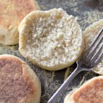 A split Homemade English Muffin with a fork beside it, surrounded by other whole English muffins on a surface dusted with cornmeal.