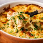 A spoon scoops a portion of cheesy, baked pasta topped with fresh chopped parsley from a ceramic dish—reminiscent of the creamy richness found in classic Hot Crab Dip.
