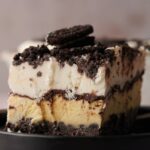 A slice of Oreo Ice Cream Cake, topped with crumbled cookies, sits on a black plate.
