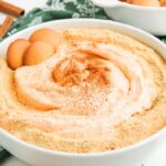 A bowl of creamy Pumpkin Pie Dip topped with crushed cookies, cinnamon, and three whole vanilla wafers, set on a green patterned cloth.