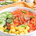 A bowl of rice topped with grilled spicy salmon, sliced cucumber, shredded carrots, diced mango, jalapeño slices, green onions, and sesame seeds—perfect for fans of Spicy Salmon Bowls.