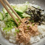 A Spicy Tuna Bowl featuring white rice topped with sliced cucumber, shredded seaweed, chopped green onions, sesame seeds, and a portion of tuna salad, served with chopsticks resting on the bowl.