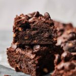 Two thick Sweet Potato Brownies are stacked on top of each other, topped with chocolate chips, with more brownies blurred in the background.