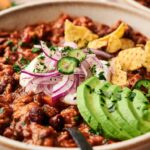 A bowl of turkey chili topped with sliced avocado, jalapeño, red onion, sour cream, chopped herbs, and tortilla chips.