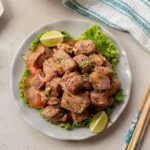 A plate of Vietnamese Shaking Beef features cooked beef cubes with onions and tomato slices, served on lettuce leaves and garnished with lime wedges. Chopsticks and a napkin are placed beside the plate.