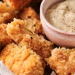 Close-up of crispy zucchini fries next to a bowl of creamy dipping sauce, with a lemon wedge partially visible.