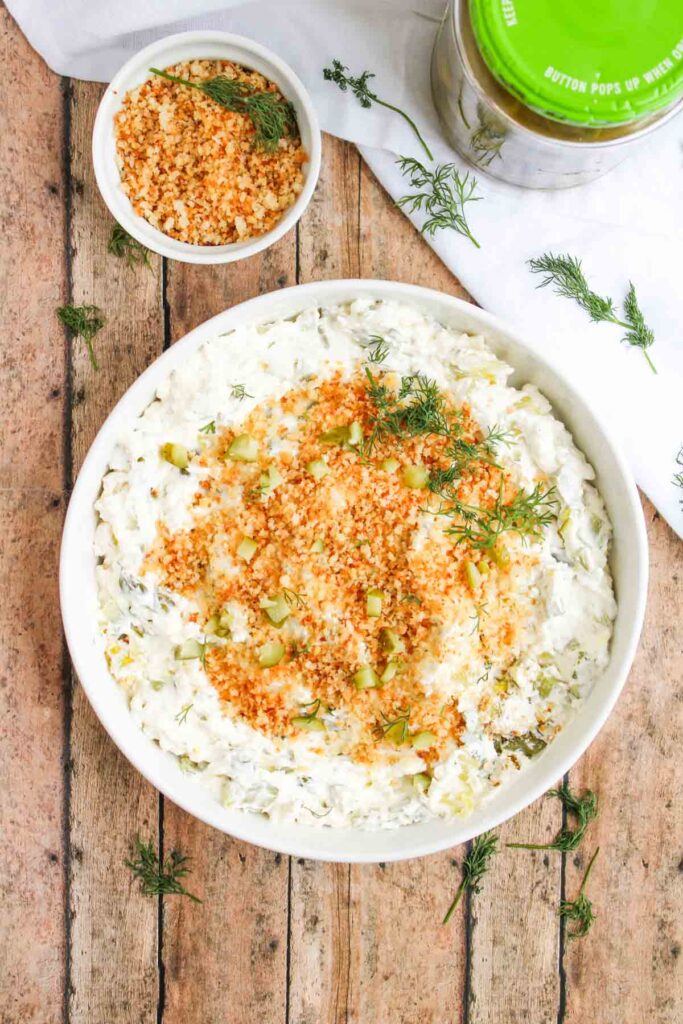 A bowl of creamy Fried Dill Pickle Dip topped with breadcrumbs, chopped pickles, and fresh dill sits on a wooden surface beside a small dish of breadcrumbs and a jar of pickles.