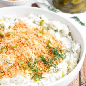 A white bowl filled with creamy Fried Dill Pickle Dip, topped with breadcrumbs, diced pickles, and fresh dill sits on a wooden surface. A small dish and a jar of pickles are in the background.