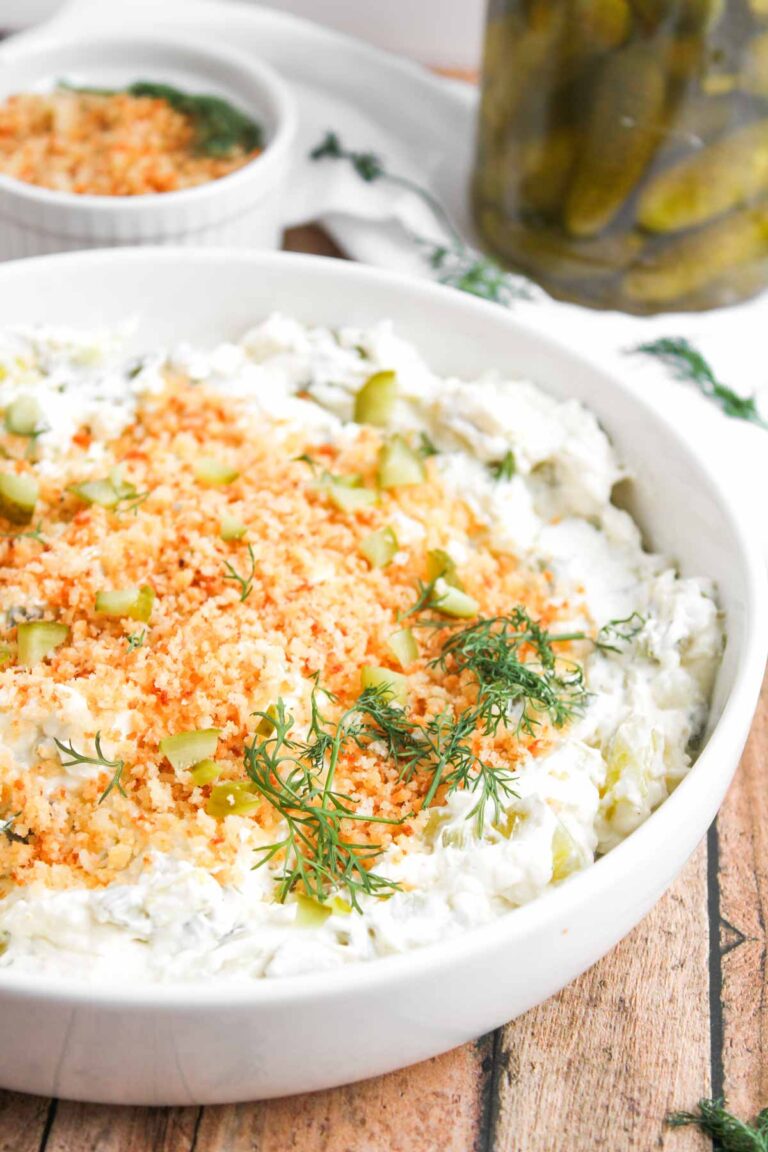 A white bowl filled with creamy Fried Dill Pickle Dip, topped with breadcrumbs, diced pickles, and fresh dill sits on a wooden surface. A small dish and a jar of pickles are in the background.