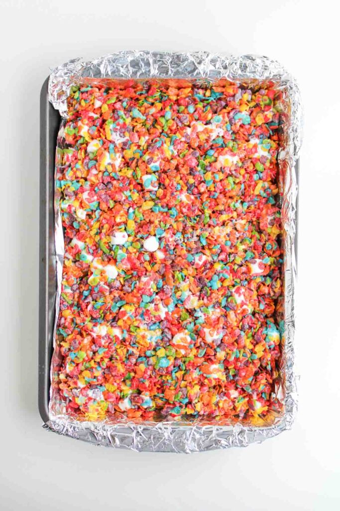 A baking tray lined with foil holds a large, rectangular sheet of colorful Fruity Pebble Rice Krispies treats made with marshmallows.