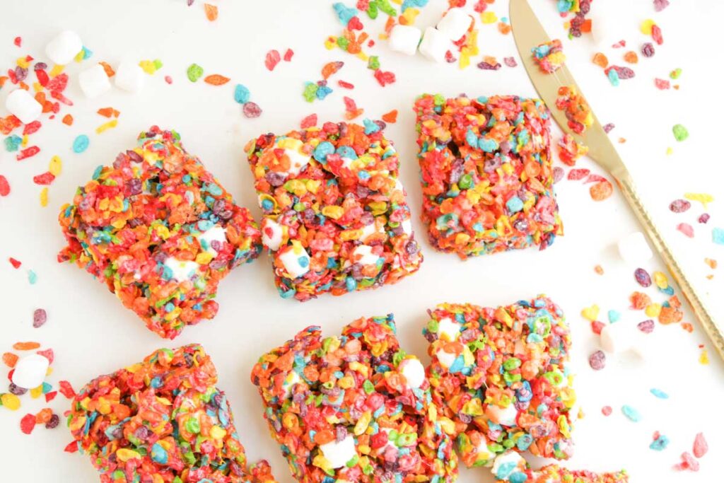 Six squares of Fruity Pebble Rice Krispies marshmallow treats are arranged on a white surface, surrounded by scattered cereal pieces, mini marshmallows, and a gold knife.