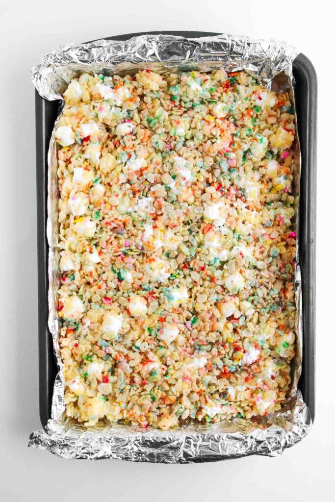 A baking pan lined with foil contains colorful Funfetti Rice Krispie Treats, with visible melted marshmallows and rainbow sprinkles.