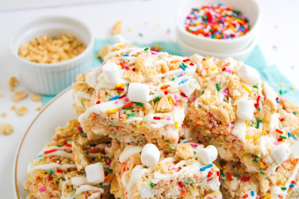 A plate of Funfetti Rice Krispie Treats topped with white icing, mini marshmallows, and colorful sprinkles. Small bowls of cereal and sprinkles are in the background.