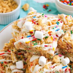 A plate of Funfetti Rice Krispie Treats topped with mini marshmallows, colorful sprinkles, and white drizzle. Bowls of sprinkles and cereal are visible in the background.