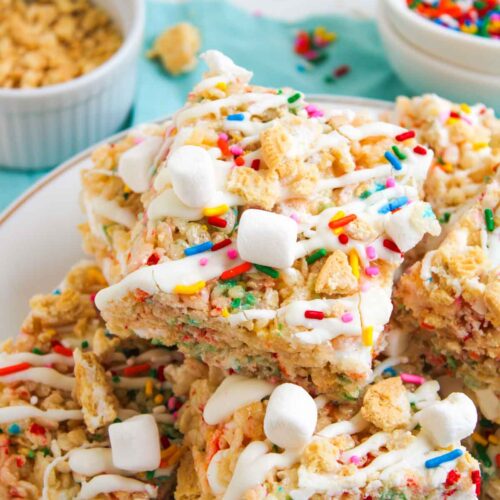 A plate of Funfetti Rice Krispie Treats topped with mini marshmallows, colorful sprinkles, and white drizzle. Bowls of sprinkles and cereal are visible in the background.