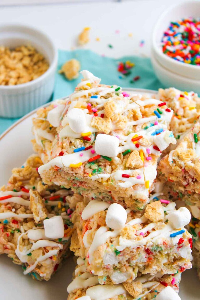 A plate of Funfetti Rice Krispie Treats topped with mini marshmallows, colorful sprinkles, and white drizzle. Bowls of sprinkles and cereal are visible in the background.