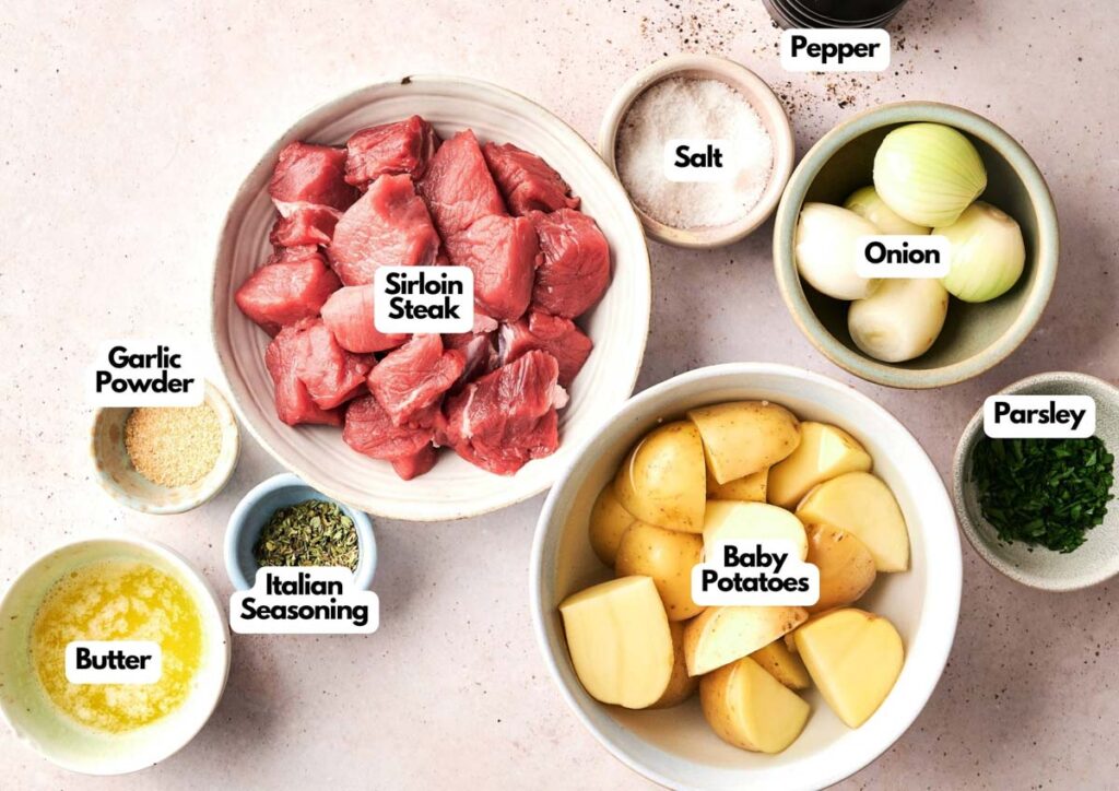 Bowls of Garlic Butter Steak and Potatoes Foil Packet ingredients: sirloin steak cubes, halved baby potatoes, chopped onions, parsley, melted butter, Italian seasoning, garlic powder, salt, and pepper on a light surface.