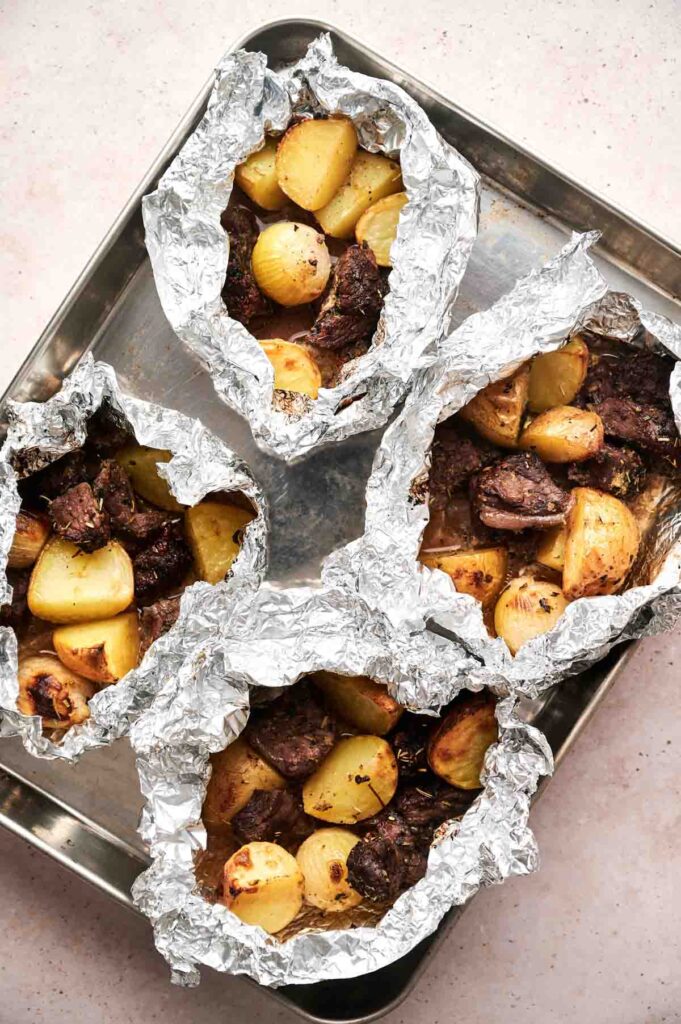 Four Garlic Butter Steak and Potatoes Foil Packets filled with roasted potatoes and tender beef are arranged on a metal baking tray.