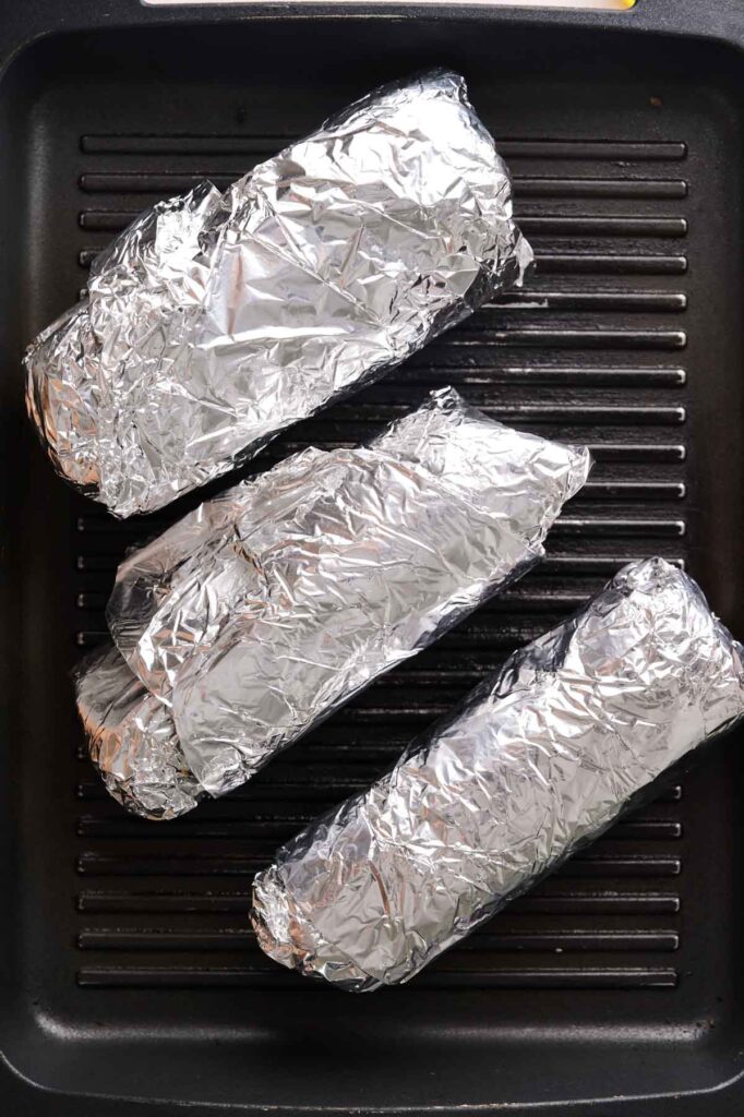 Three foil-wrapped Grilled Cheese Burritos are placed on a grill pan with ridges.