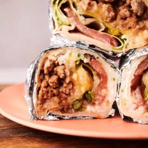 Three foil-wrapped Grilled Cheese Burritos cut in half, stacked on a peach-colored plate, reveal layers of ground meat, cheese, lettuce, and tomato.
