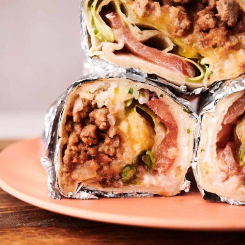Three foil-wrapped Grilled Cheese Burritos cut in half, stacked on a peach-colored plate, reveal layers of ground meat, cheese, lettuce, and tomato.