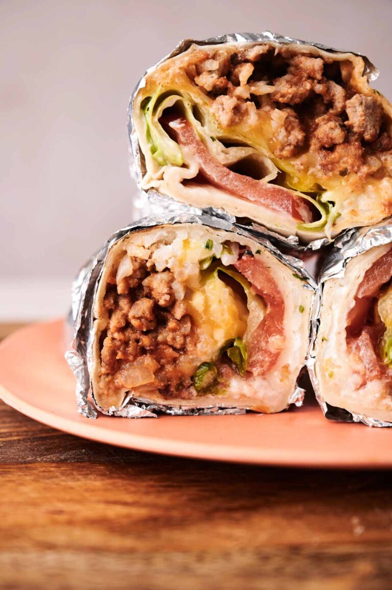 Three foil-wrapped Grilled Cheese Burritos cut in half, stacked on a peach-colored plate, reveal layers of ground meat, cheese, lettuce, and tomato.