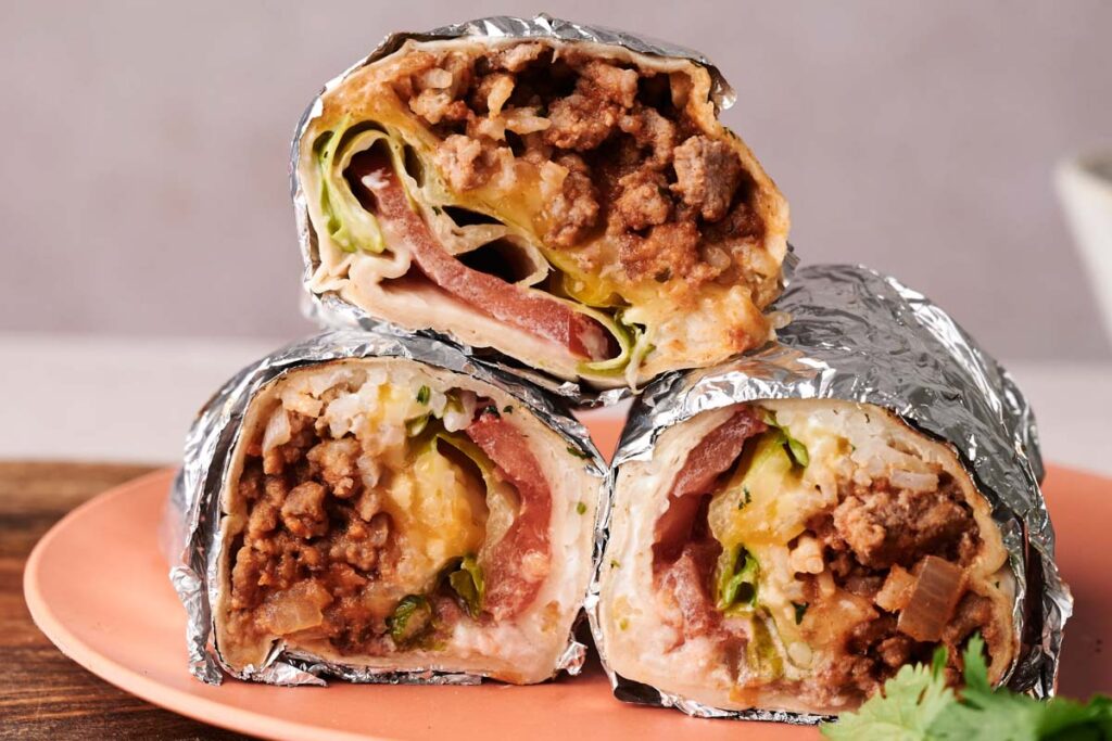 Three halved Grilled Cheese Burritos wrapped in foil, showing ground meat, cheese, lettuce, tomato, and rice filling, stacked on a pink plate.