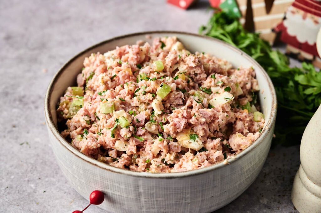 A bowl of Ham Salad, featuring chopped ham, celery, and fresh herbs, sits on a gray surface next to sprigs of fresh parsley.
