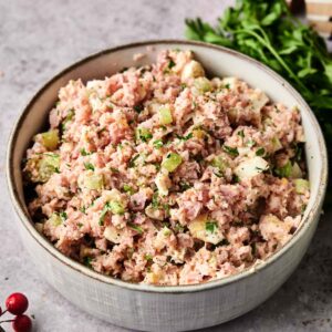 A bowl of homemade Ham Salad featuring diced ham, celery, pickles, and herbs sits on a gray countertop, garnished with parsley and surrounded by festive decorations.
