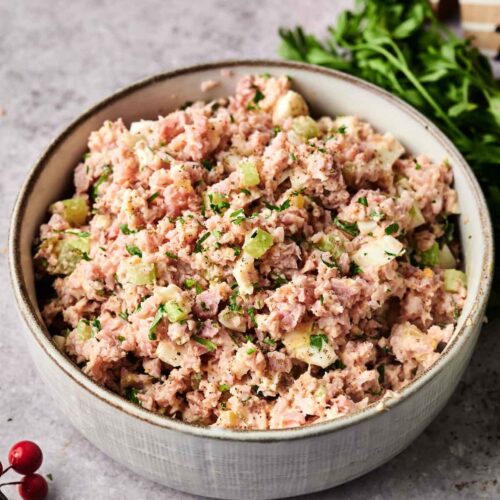 A bowl of homemade Ham Salad featuring diced ham, celery, pickles, and herbs sits on a gray countertop, garnished with parsley and surrounded by festive decorations.