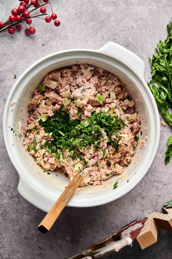 A white mixing bowl filled with creamy ham salad, chopped herbs on top, and a wooden spoon, with parsley and red berries nearby on a gray surface.