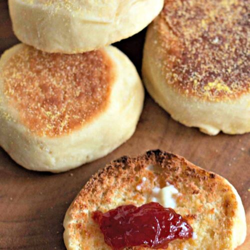 A group of Homemade English Muffins, with one split open and topped with butter and strawberry jam, displayed on a wooden surface.