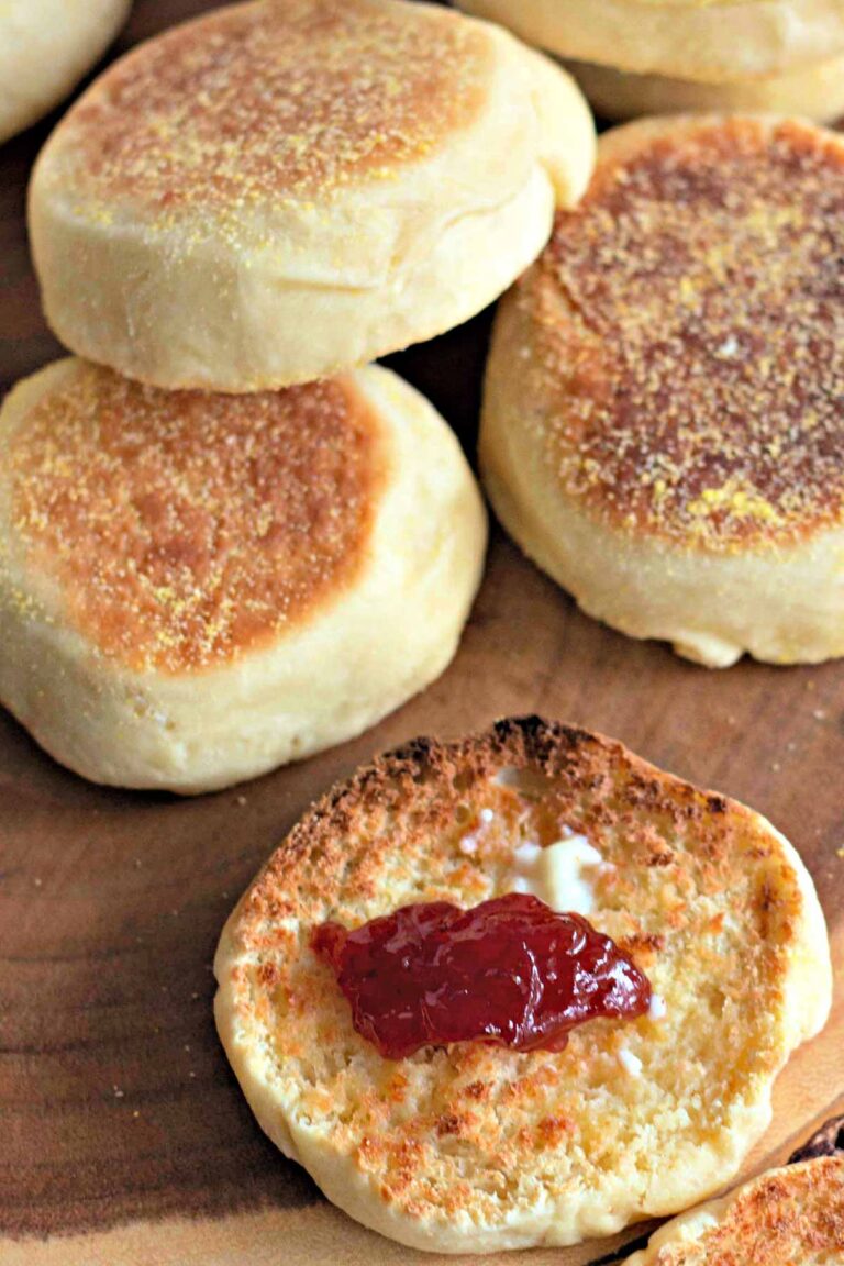 A group of Homemade English Muffins, with one split open and topped with butter and strawberry jam, displayed on a wooden surface.