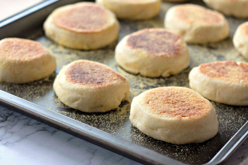A baking sheet with several freshly baked homemade English muffins, lightly browned on top and sprinkled with cornmeal, sits on a marble countertop.