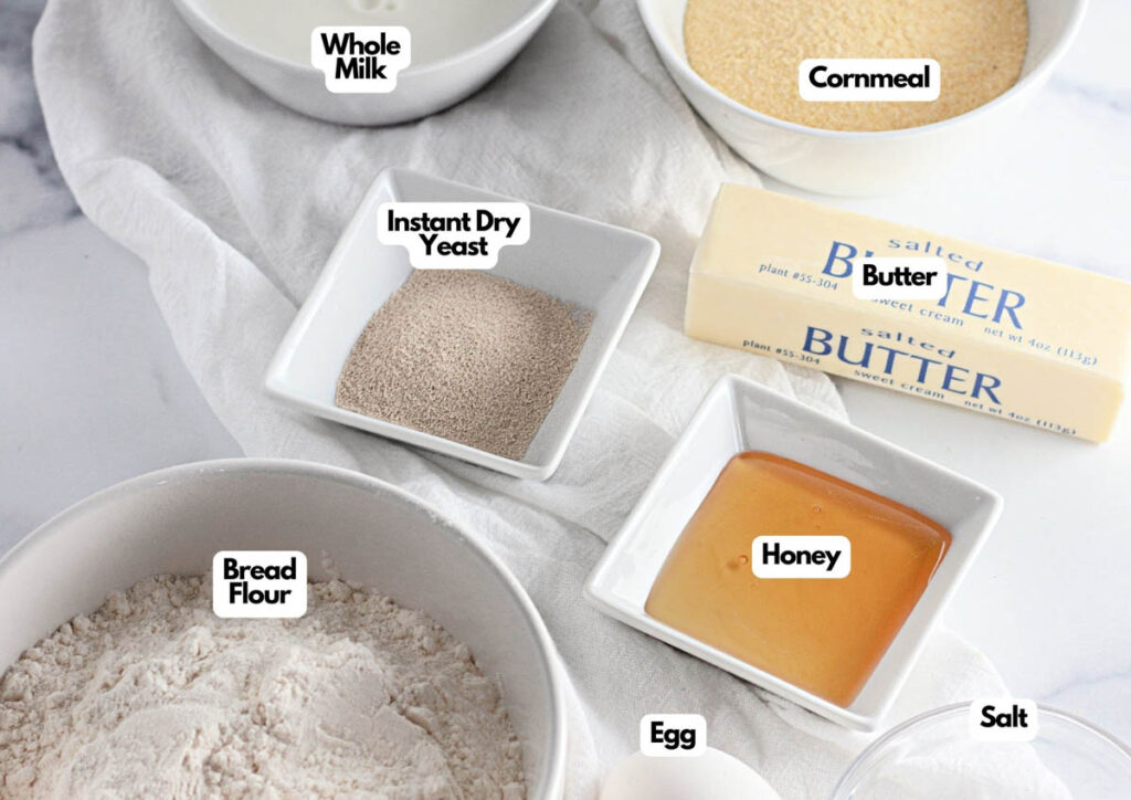 Ingredients for Homemade English Muffins are arranged on a table, including whole milk, cornmeal, instant dry yeast, butter, bread flour, honey, an egg, and salt.